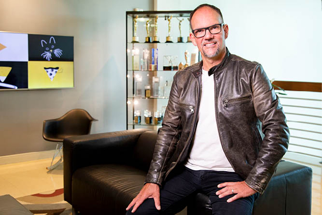 A smiling man sitting on the arm of a brown couch