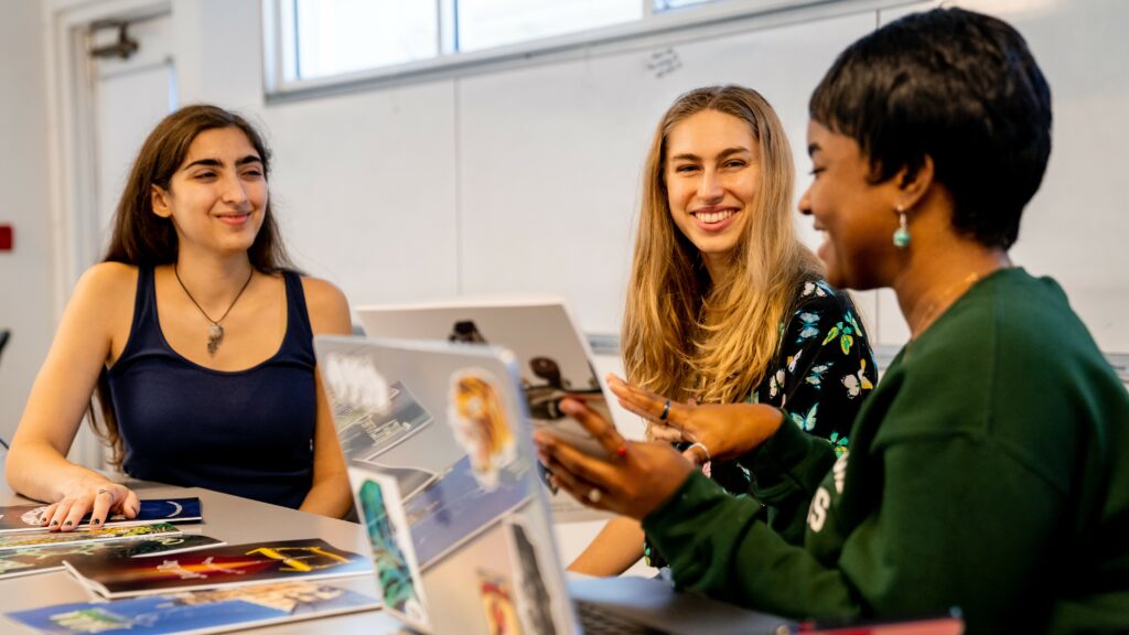 Three female college students work collaboratively, sharing a laptop and printed zines at a white table.