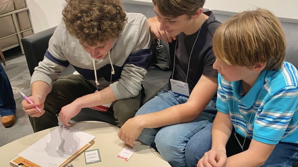 Three young people sit on a black couch and play a table-top game with dice, playing cards, with a writing component.