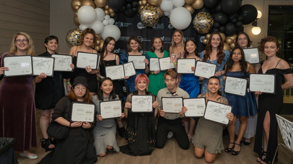 Seventeen people pose in front of a display with gold, black, and white balloons, holding awards.