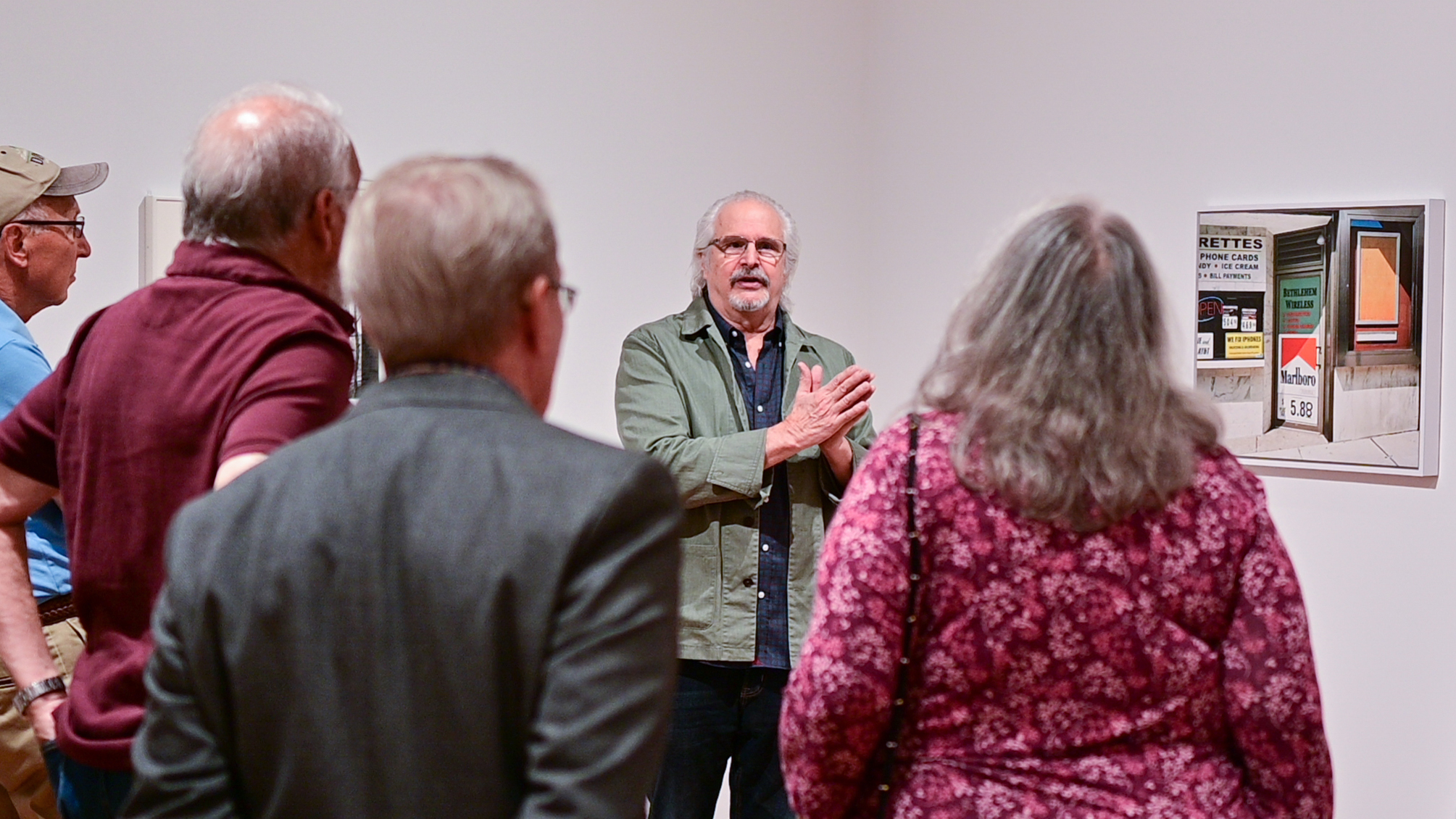 A man standing in front of a color photograph in a museum giving a talk to a group of people.