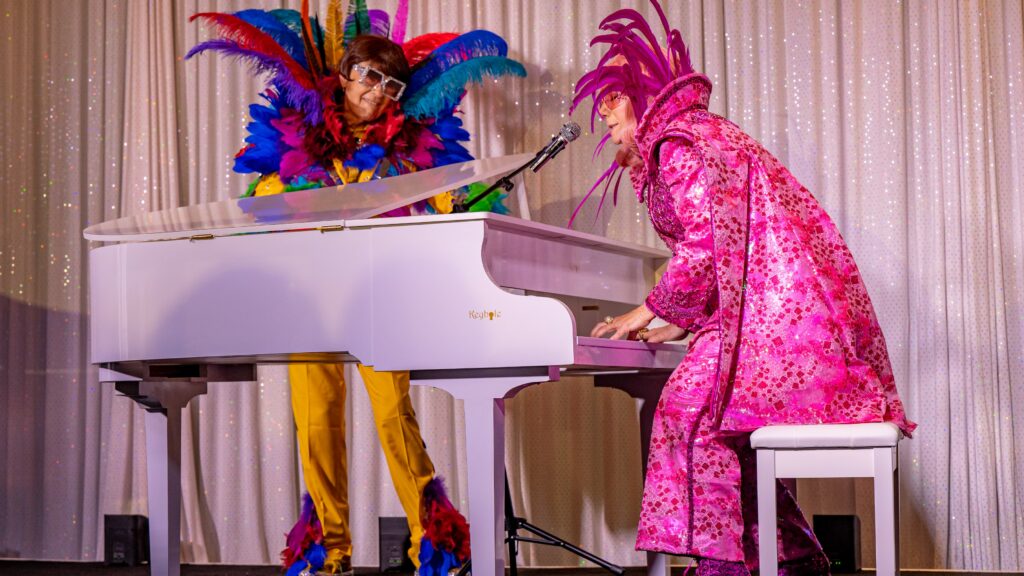 Woman with pink shimmery costume performs at piano while brightly costumed man looks on.