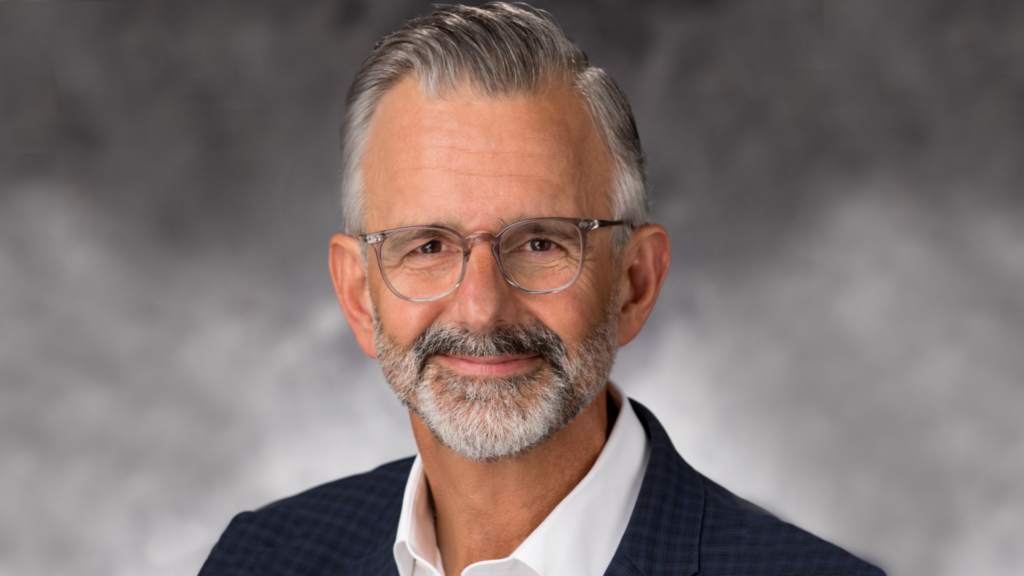 Professional headshot of a man with a beard and glasses against a gray background.