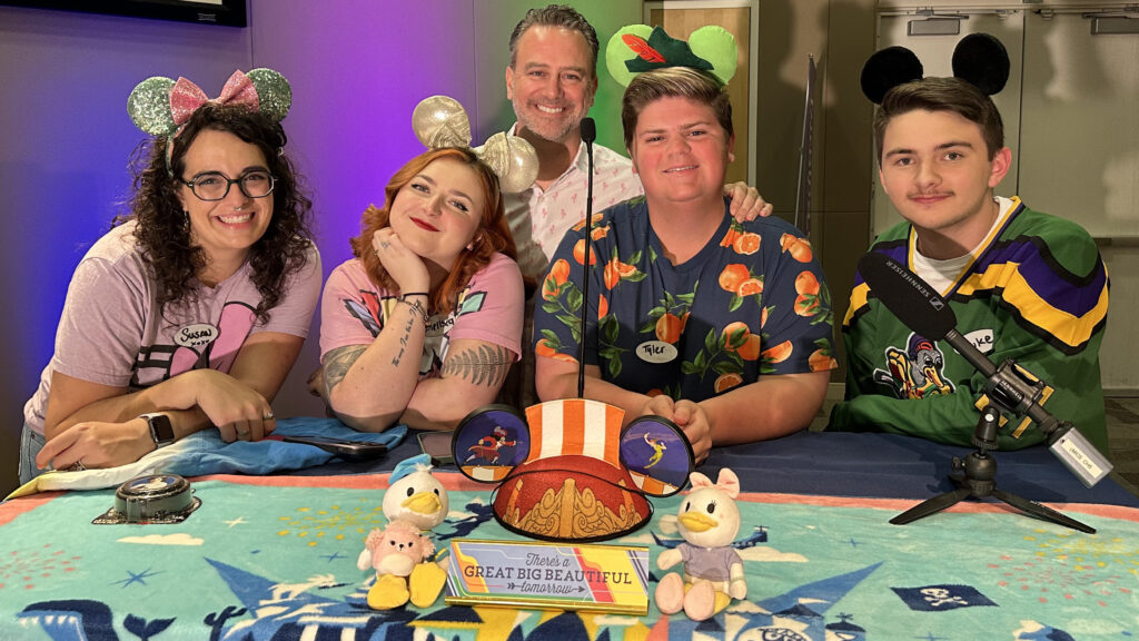 Five people pose with mouse ears while leaning on a table with Disney themed objects.