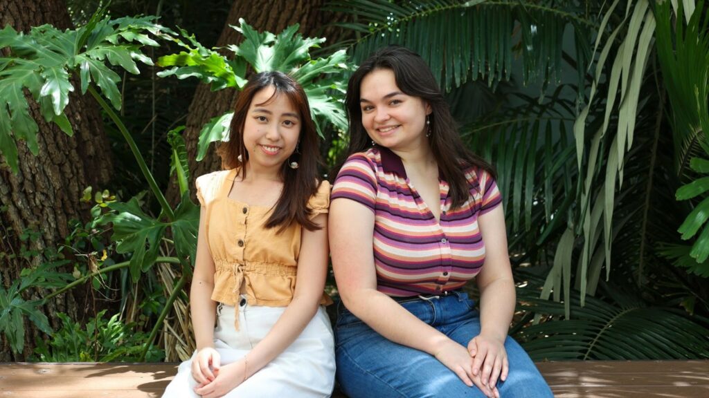 Two female college students sit back to back on a bench in front of a green foliage backdrop