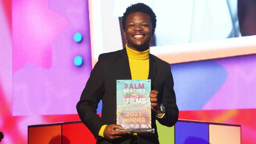 A Black male college student wearing a yellow shirt and black jacket holds up an award on stage against a colorful background