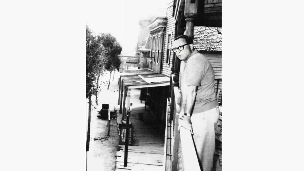 A black and white image of a man leaning on a balcony overlooking a western movie film set.