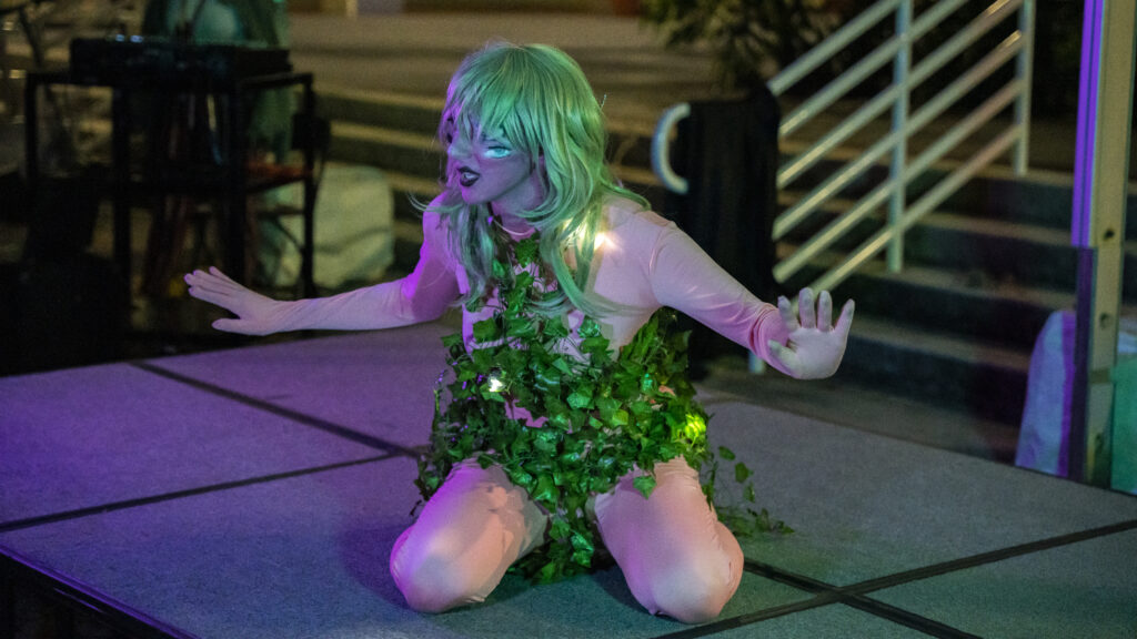 Drag performer with surrealesque large eyes and extra eyes on cheeks, covered with viney leaves.