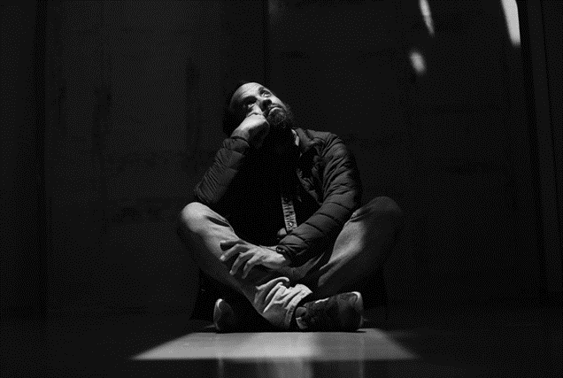 Black and white photo of a man sitting cross-legged and looking up with a spotlight on him.