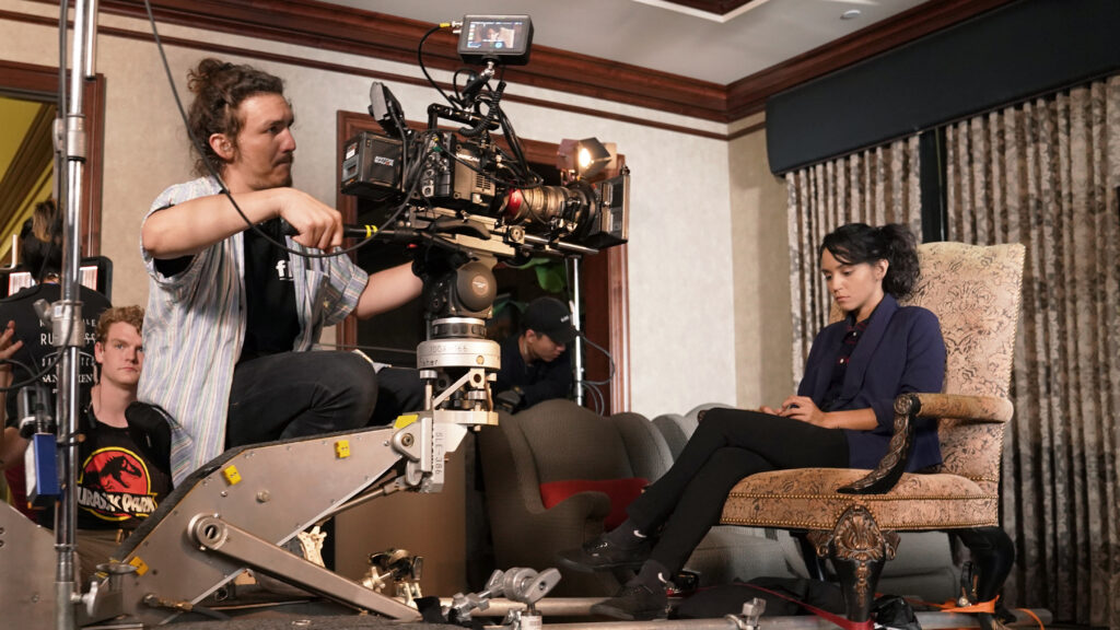 A man points a film camera at a girl sitting in an armchair inside an old house on a movie set.