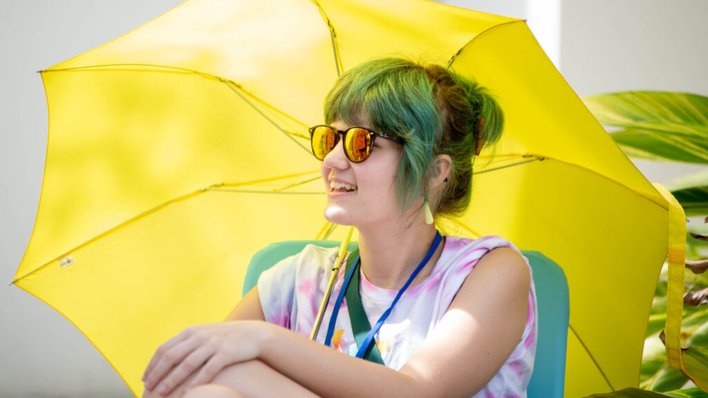 A female college student smiles under a large yellow umbrella wearing sunglasses.