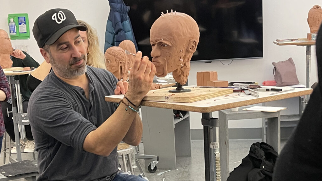 Man wearing a black baseball cap and jeans works on a clay sculpture of a human head and face