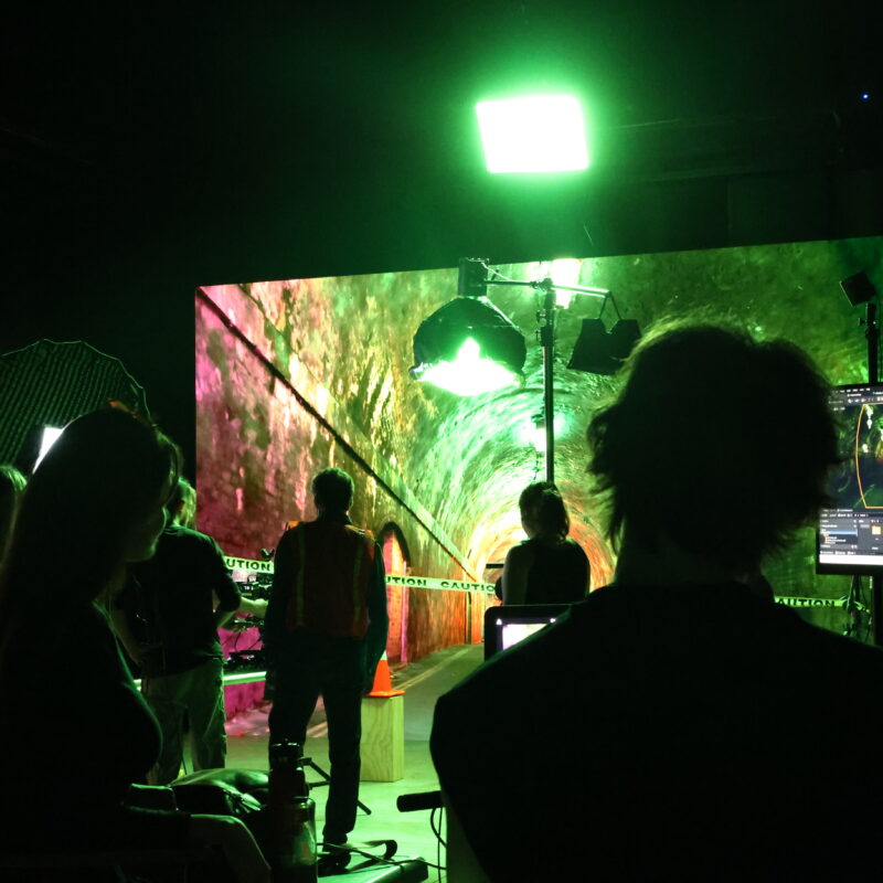 Silhouettes of people filming and working on a dark set in front of an LED screen displaying a green and red tunnel.