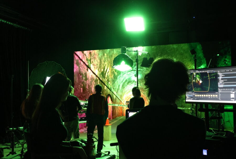 Silhouettes of people filming and working on a dark set in front of an LED screen displaying a green and red tunnel.