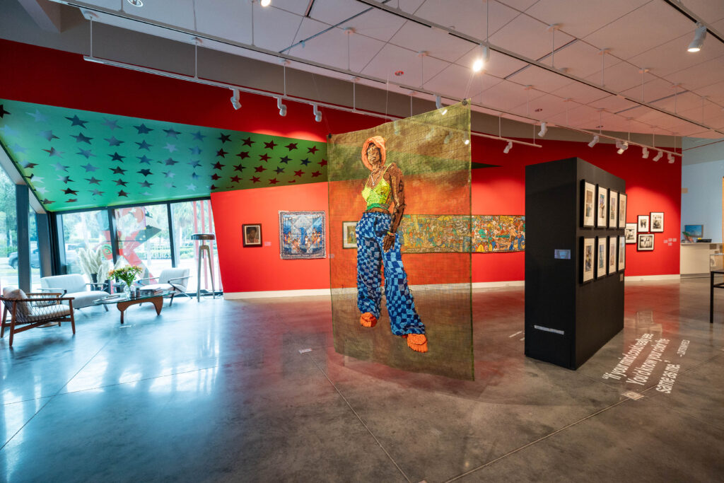 Installation view of a gallery with red walls and a green wall covered in stars, with a large figurative textile artwork hanging in the middle of the gallery.