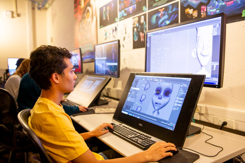 Student uses a drawing tablet and computer to create a digital character design in a classroom setting.
