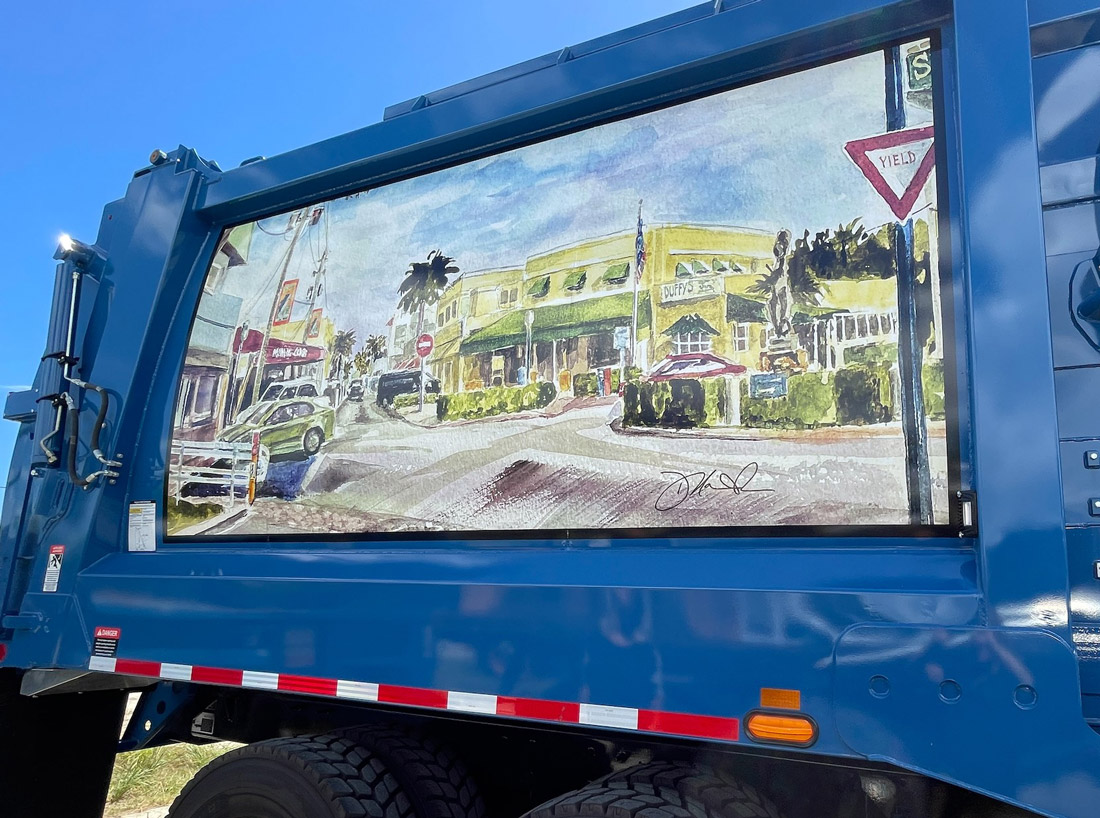 Close-up of a painted street scene mural on a blue truck, depicting colorful buildings and a sunny neighborhood intersection