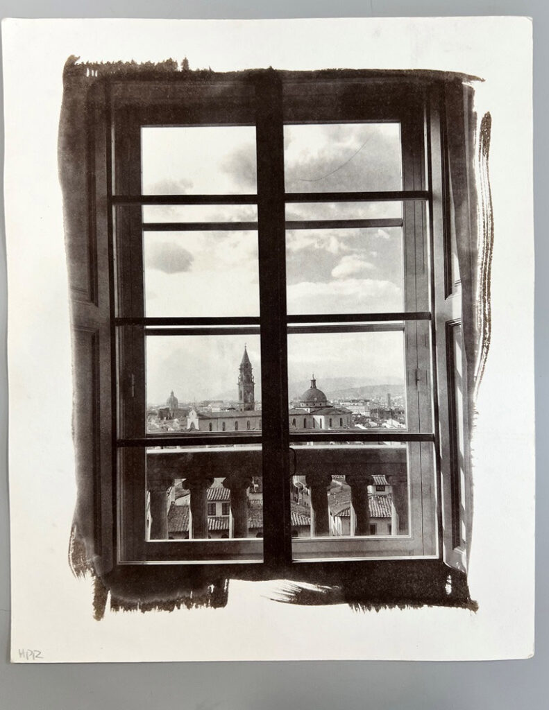 Black-and-white photographic print of a window overlooking a cityscape with architectural details framed by brush-like edges
