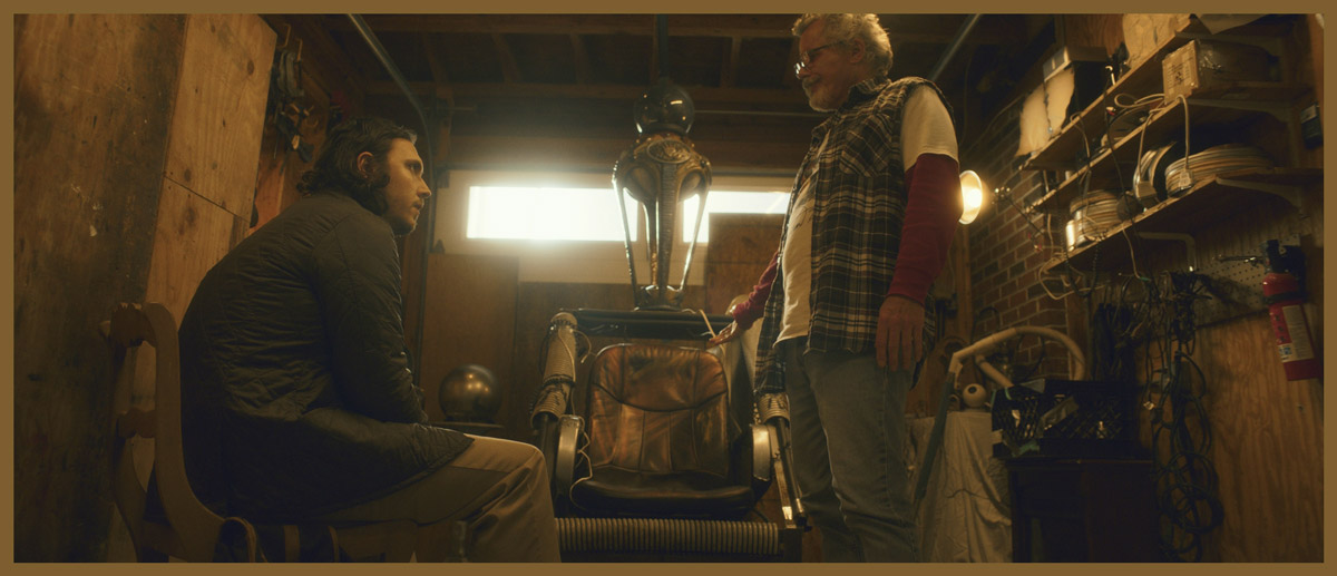 Film still of two men in a dimly lit workshop, one seated and one standing, beside a mechanical chair or device surrounded by tools and equipment