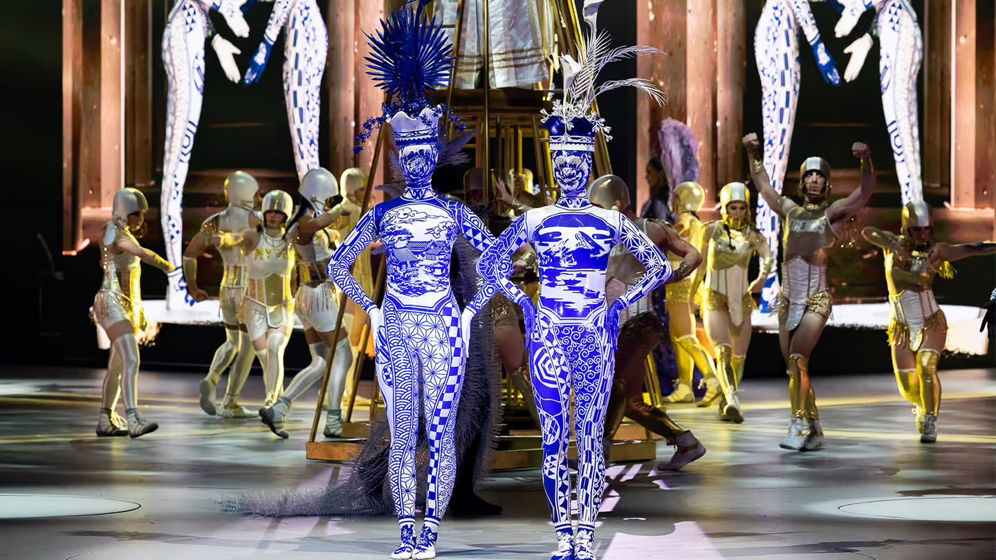 Stage performance featuring two models in blue-and-white patterned bodysuits with elaborate headpieces, surrounded by dancers in gold costumes