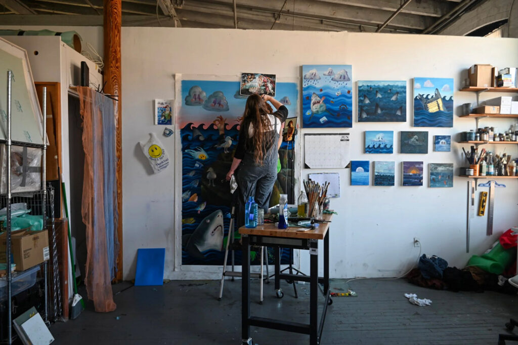 Artist working in a studio surrounded by paintings, tools, and reference images pinned to the wall