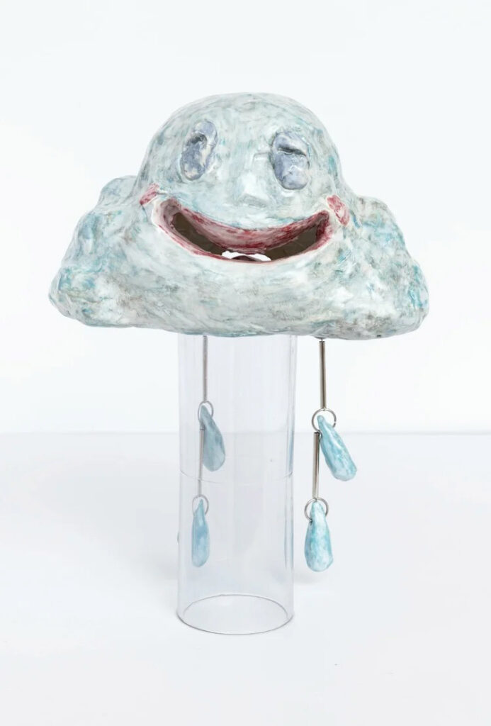 Ceramic cloud sculpture with a smiling face and dangling teardrop elements displayed on a clear pedestal