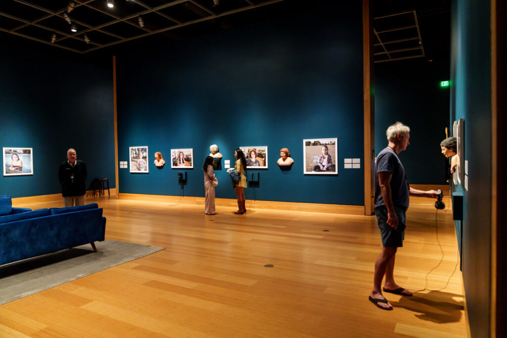 Visitors walking through a museum gallery with blue walls displaying framed photographs and sculptural portrait busts