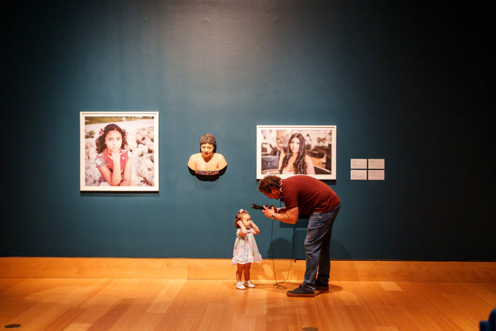 Adult interacting with a child in front of framed portrait artwork and a sculptural bust in a museum gallery