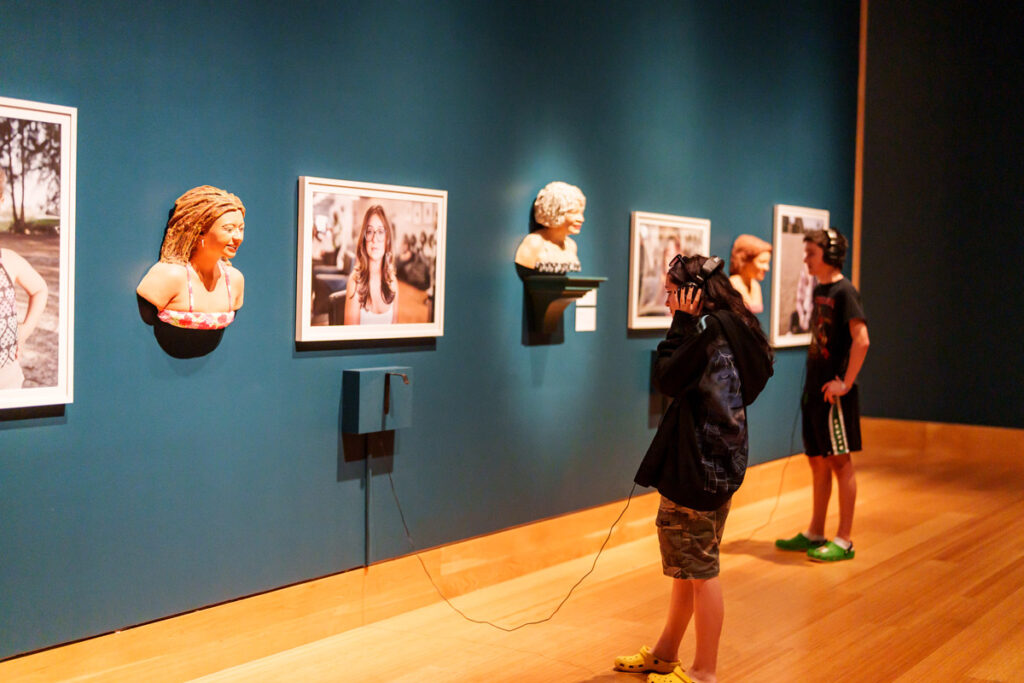 Visitor using headphones on front of a sculptural busts and framed portraits displayed along a blue gallery wall