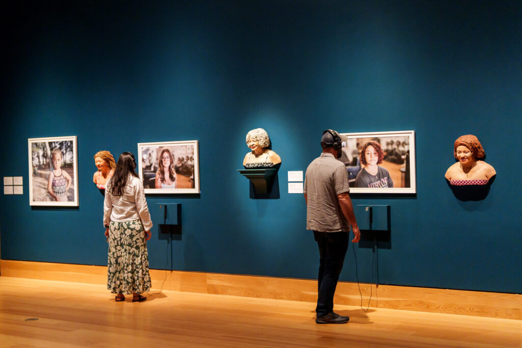 Museum visitors observing framed portrait artworks and sculptural busts arranged along a gallery wall
