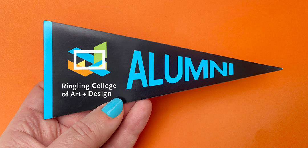 Hand holding a black triangular alumni pennant with Ringling College of Art and Design logo and teal “Alumni” text against an orange background